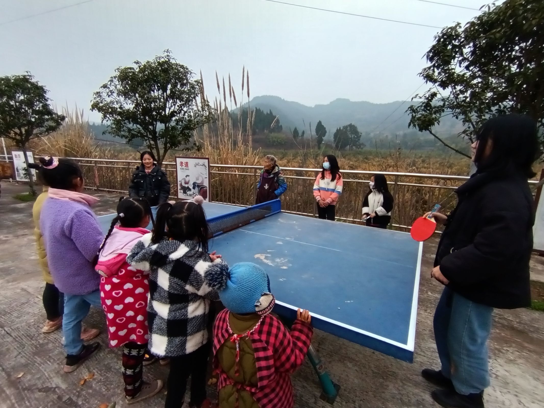 图为团队成员在李渡镇枣垭寺村童伴之家与小朋友们一起热情地玩耍活动。一些小朋友们在乒乓球台边驻足观看， ...